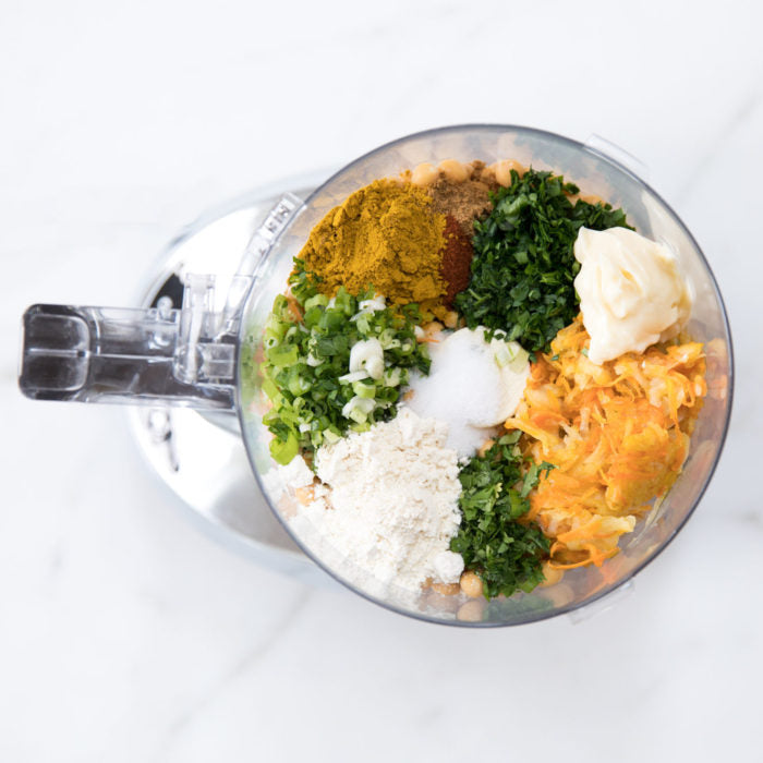 Top-down food processor with chickpea burger ingredients: chickpeas, shredded potato, scallions, parsley, curry powder, flour, and mayonnaise