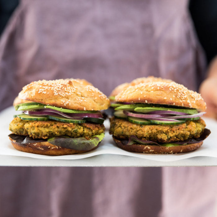Front view of two chickpea burgers with golden patties, cucumber, red onion, and sesame buns against a blurred mauve apron