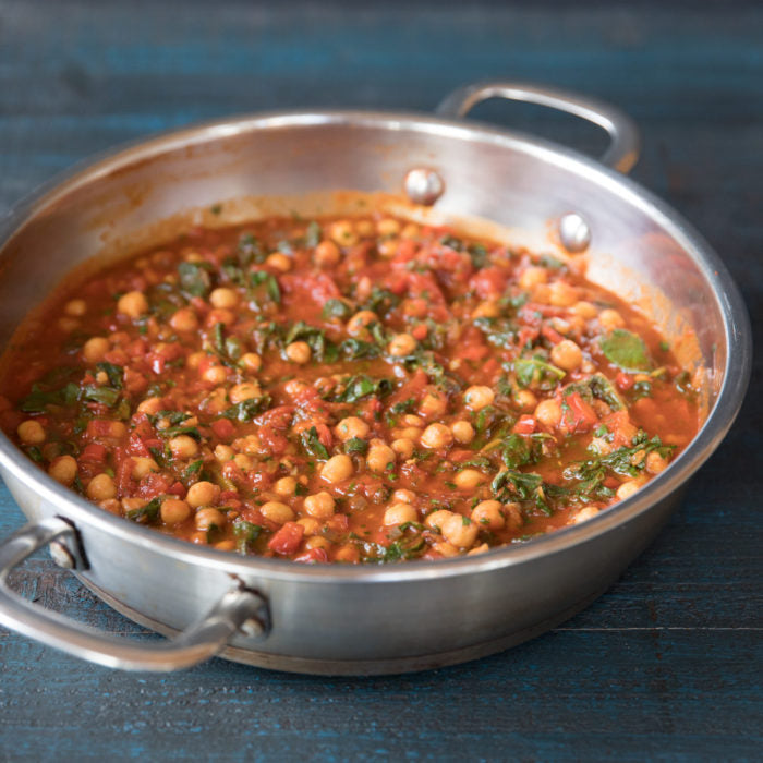 Overhead view of chickpea and tomato stew in a stainless-steel skillet with wilted greens on dark wood