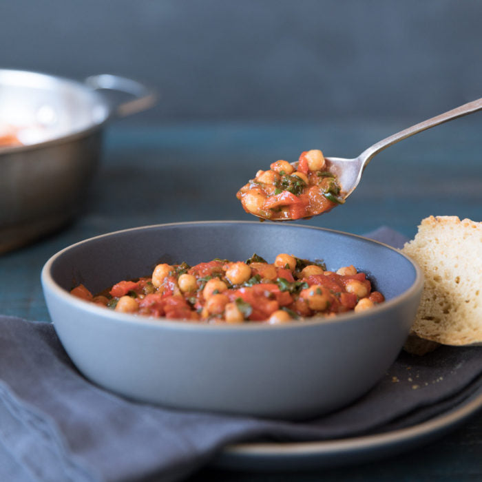 Spoon lifting chickpea and tomato stew from a blue bowl beside a slice of crusty bread on a dark teal table