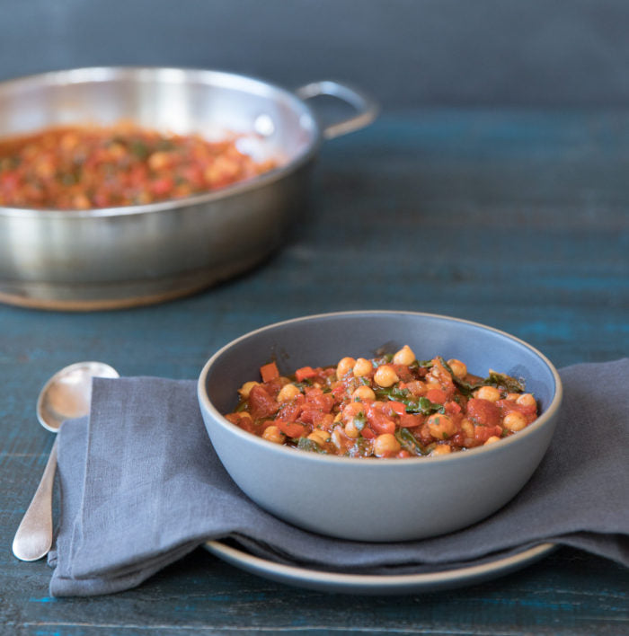 Chickpea and tomato stew served in a blue bowl on a folded napkin, spoon beside it, with a skillet of stew blurred in the background