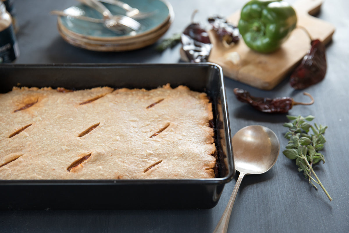 Einkorn chili con carne pot pie in a 9 x 13-inch baking pan on a dark tabletop with a serving spoon, poblano pepper, dried chiles, and fresh herbs nearby