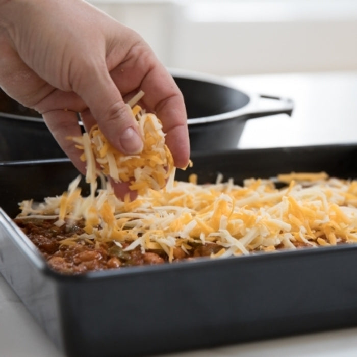 Hand sprinkling shredded cheddar over Einkorn chili con carne filling in a rectangular baking dish