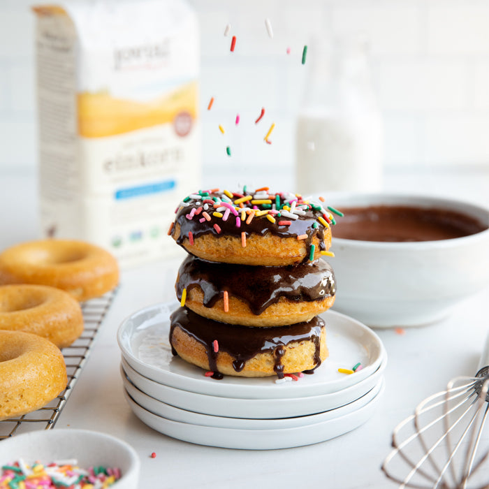 Three baked einkorn donuts stacked with chocolate glaze and falling rainbow sprinkles, bowl of chocolate and Jovial einkorn flour bag in the background