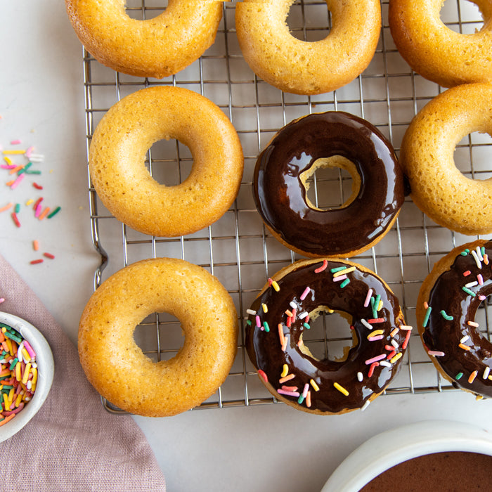 Overhead view of baked Einkorn donuts on a cooling rack; three chocolate glazed, two with rainbow sprinkles