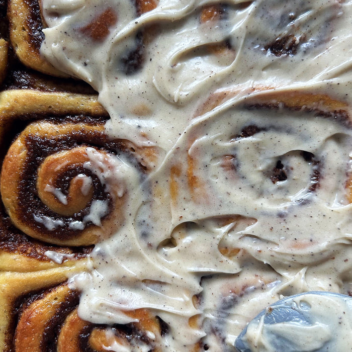 Close-up top view of sourdough cinnamon rolls with swirls of cinnamon and a spoon spreading speckled cream cheese frosting