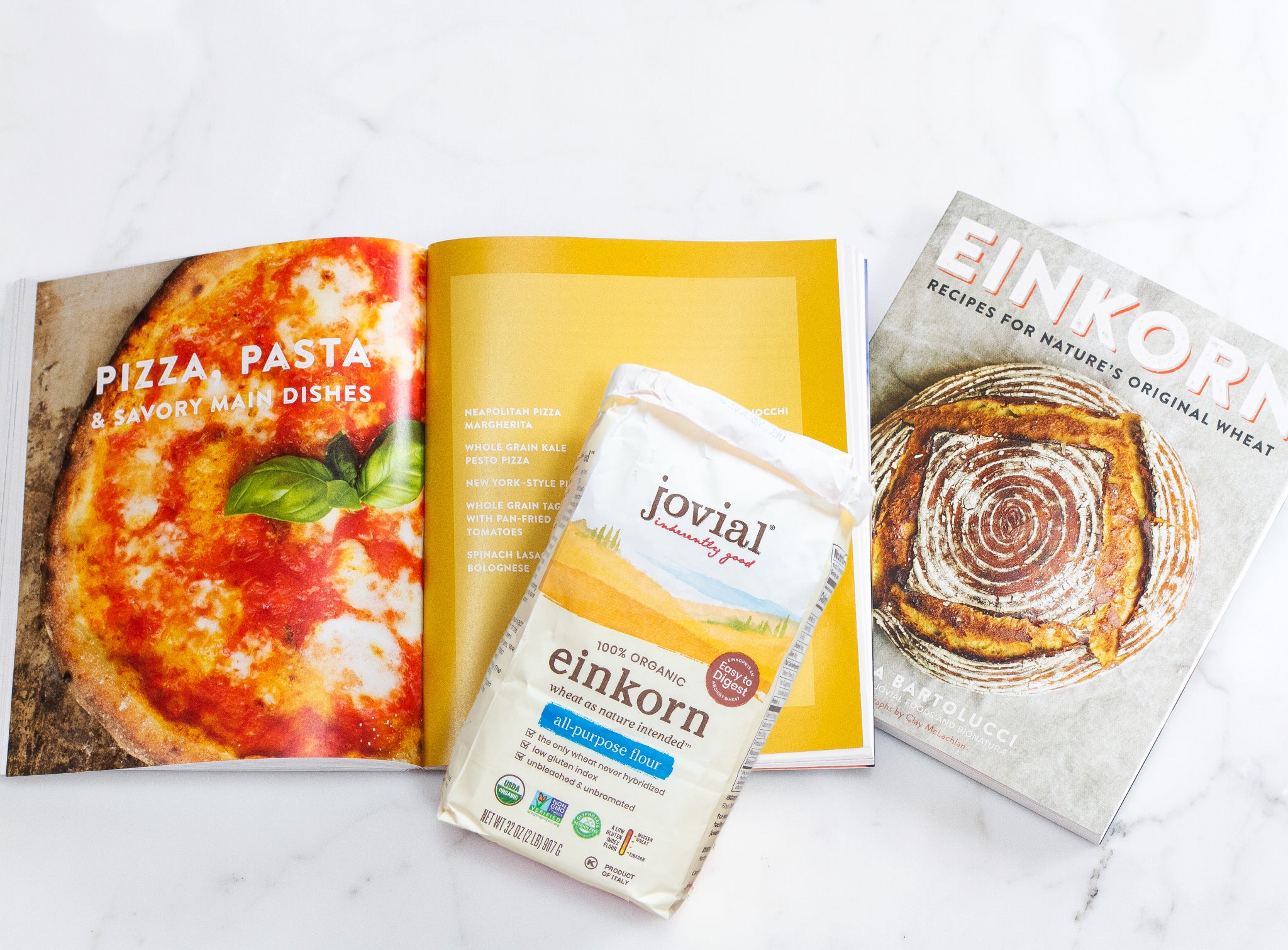 Front view of Jovial 32 oz organic einkorn flour bag showcasing label, on open pizza cookbook and closed Einkorn recipe book in background