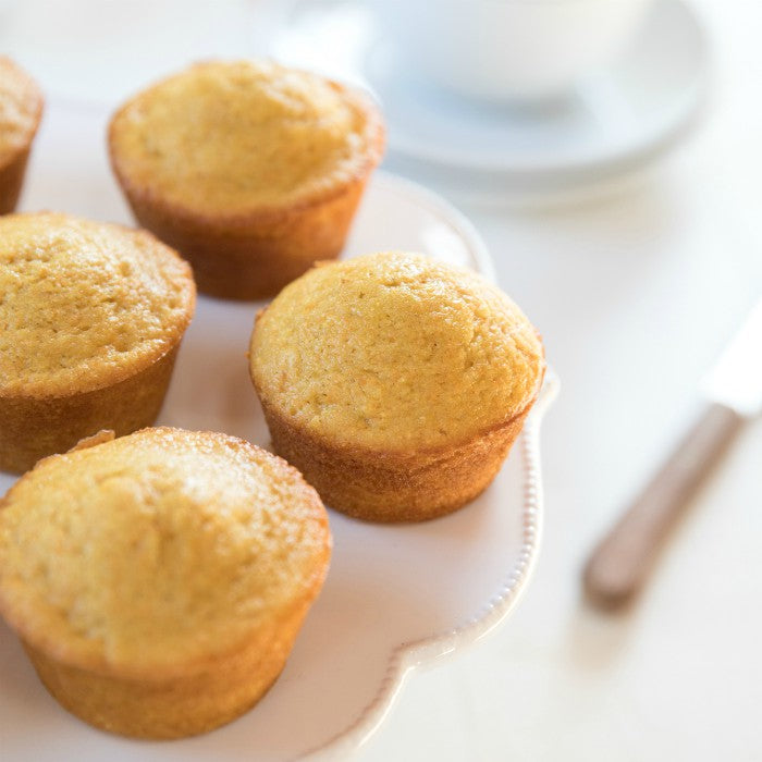 Close-up of three golden einkorn corn muffins on a white cake stand with domed tops and a blurred cup and knife behind