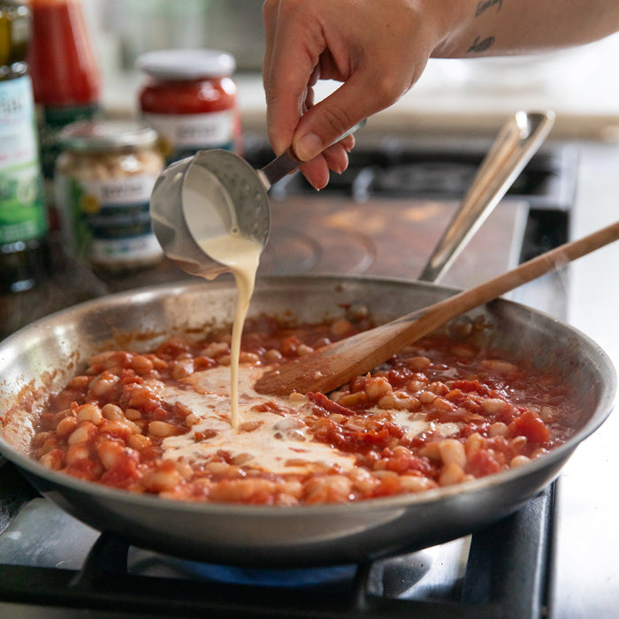 Hand pouring heavy cream into a skillet of cannellini beans and organic tomatoes on a stovetop, wooden spoon resting in pan
