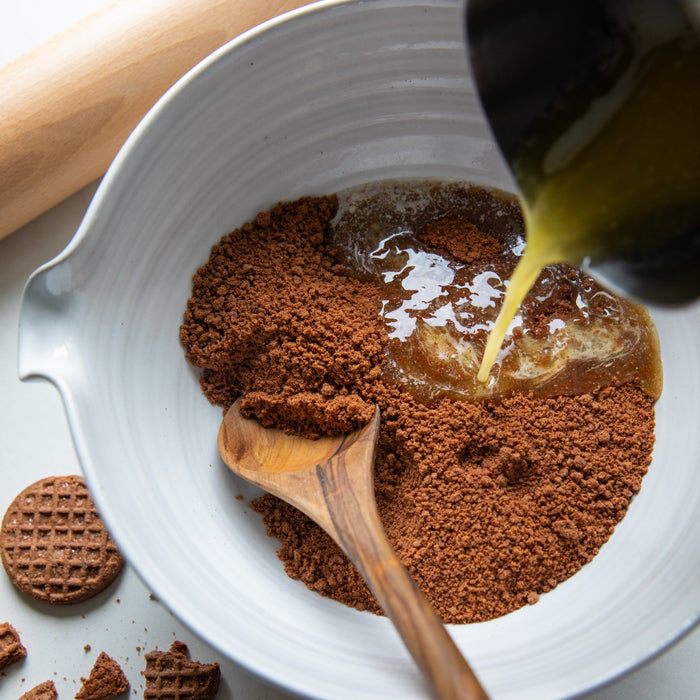 Crushed Einkorn Crispy Cocoa Cookies in a bowl with melted butter being poured for a no-bake cheesecake crust