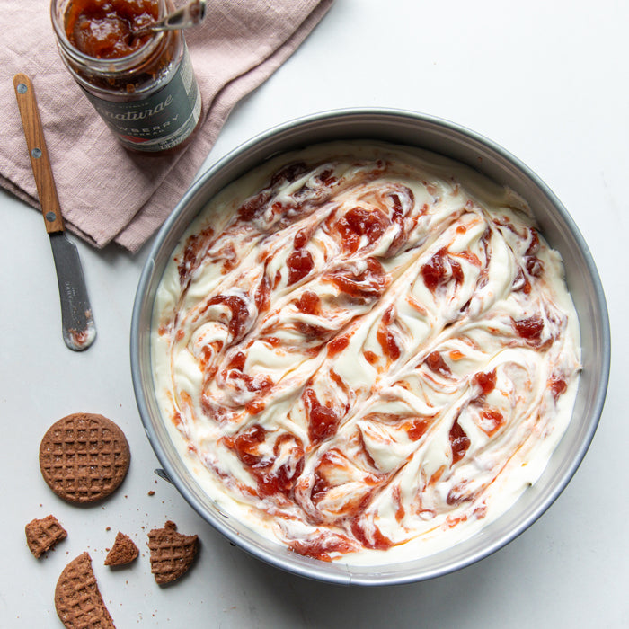 No-bake cheesecake swirled with Bionaturae strawberry fruit spread in a round pan, with crushed Einkorn Crispy Cocoa Cookies and jar nearby