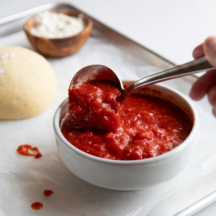 Ladle scooping chunky crushed tomato pizza sauce from a white bowl beside an einkorn pizza dough ball on a baking sheet