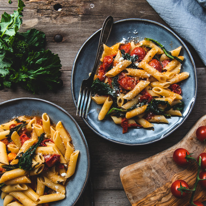 Two blue plates of penne with garlicky broccoli rabe, roasted cherry tomatoes, and grated cheese on a rustic wooden table