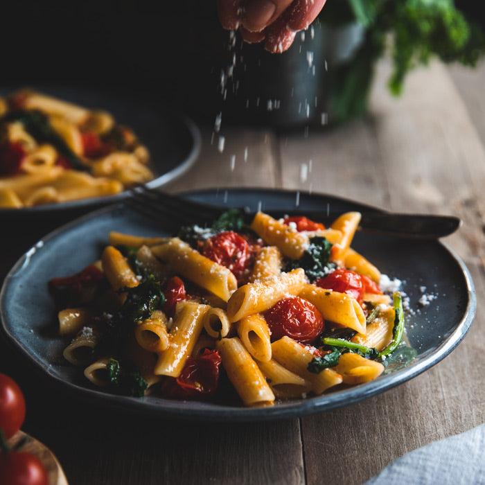 Penne pasta tossed with sautéed broccoli rabe and cherry tomatoes on a dark ceramic plate, hand sprinkling grated cheese