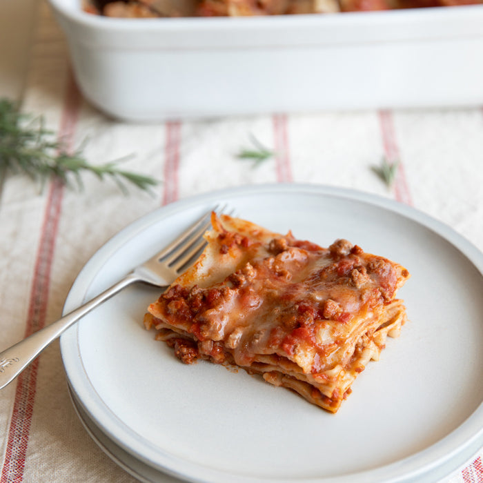 Slice of dairy-free, gluten-free lasagna Bolognese on a white plate with a fork and baking dish in the background