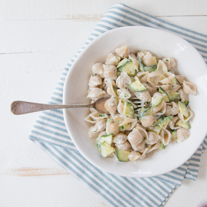 Bowl of brown rice shell pasta tossed in lemon herb Greek yogurt sauce with zucchini on a blue striped napkin