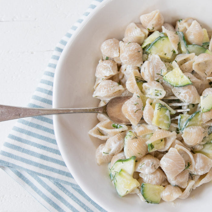Bowl of brown rice pasta shells tossed in creamy lemon‑herb Greek yogurt sauce with zucchini and dill on a striped napkin