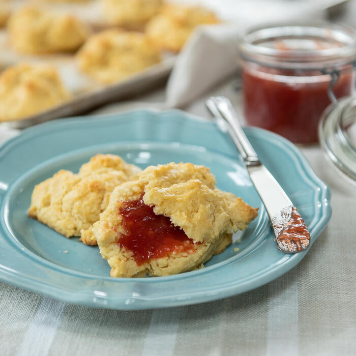 Close-up of einkorn butter drop biscuits on a blue plate, one split and spread with red jam, knife and jam jar in soft-focus background