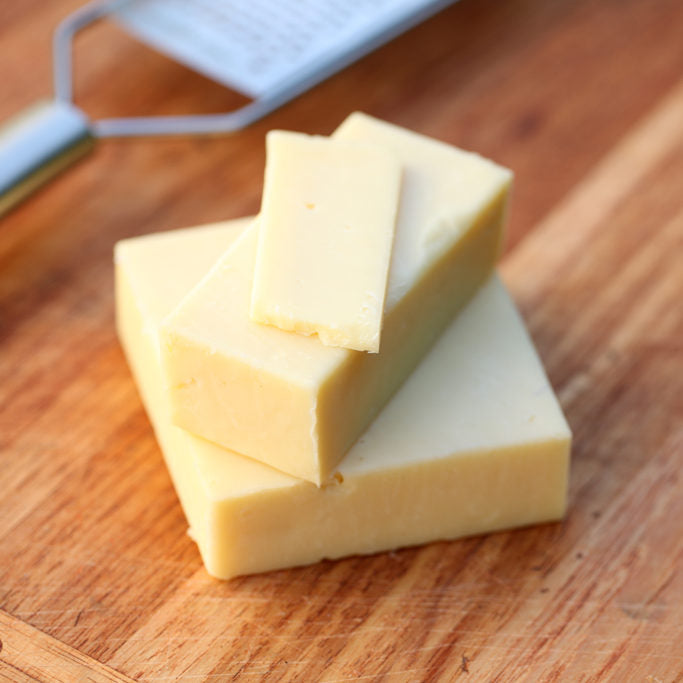 Close-up of three stacked blocks of pale yellow cheese showcasing smooth cut surfaces on a wooden cutting board with a metal grater in the background