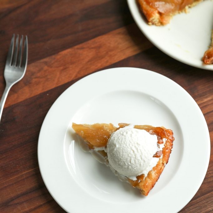 Top-down view of an Einkorn apple tarte tatin slice topped with a scoop of vanilla ice cream on a white plate over a wood table