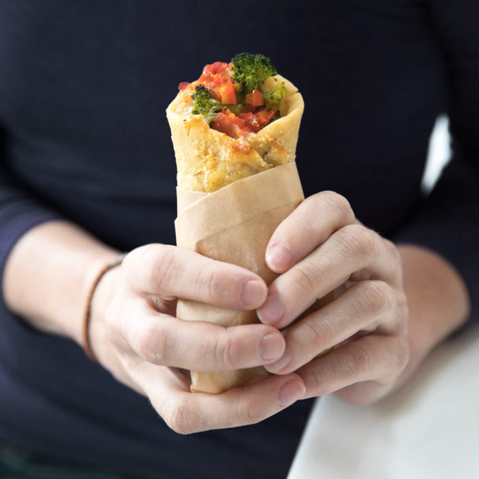 Hands holding an Einkorn pizza roll-up wrapped in parchment, filled with tomato sauce and broccoli against a dark background