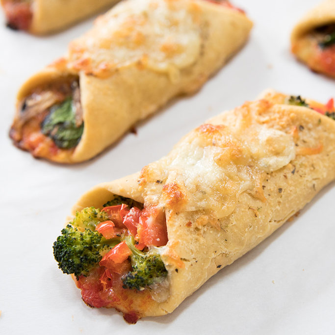 Angled close-up of einkorn pizza roll-ups with golden crust, melted cheese, and broccoli-tomato filling on parchment