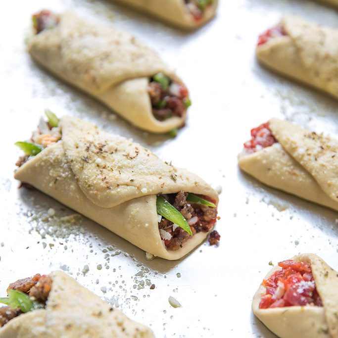 Close up of uncooked Einkorn pizza roll ups showcasing folded dough with meat and pepper filling and grated cheese on a baking sheet