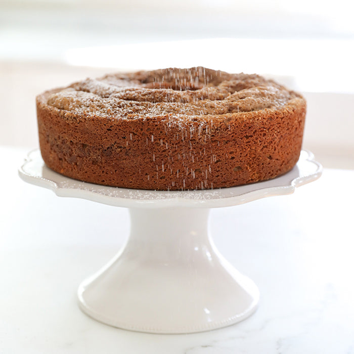 Side view of Einkorn sour cream coffee cake on a white pedestal with powdered sugar being dusted over the top