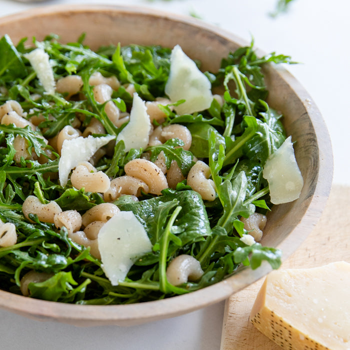 Bowl of pasta salad with arugula and shaved cheese on a light surface