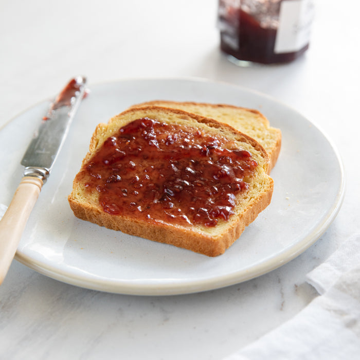 Top-down view of two slices of Einkorn sandwich bread on a white plate, one slice spread with red jam, knife and jam jar blurred in background