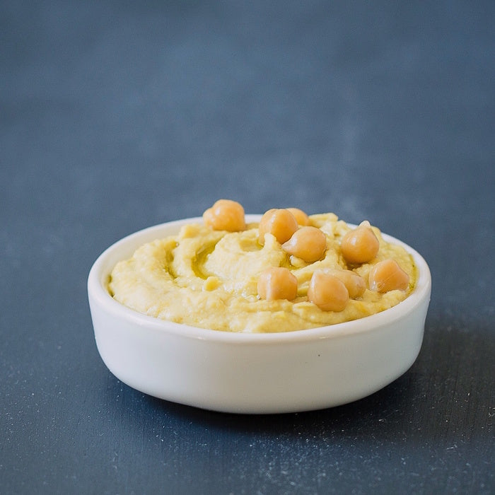Close up front view of hummus in a white bowl topped with whole chickpeas and a drizzle of olive oil on a dark slate surface