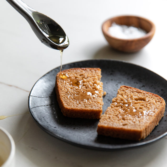 Two slices of whole-grain einkorn bread on black plate with honey drizzling over them and a wooden bowl of salt in background