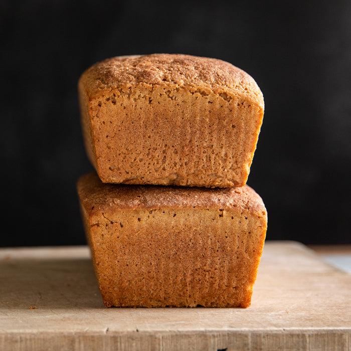 Side view of two stacked whole grain Einkorn loaves showing dense crumb and golden-brown crust on a wooden board against a dark background