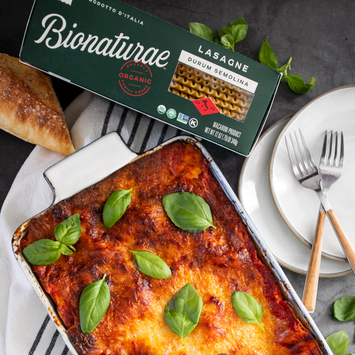 Eggplant lasagna topped with golden cheese and basil, beside a Bionaturae lasagne box, bread, and plates