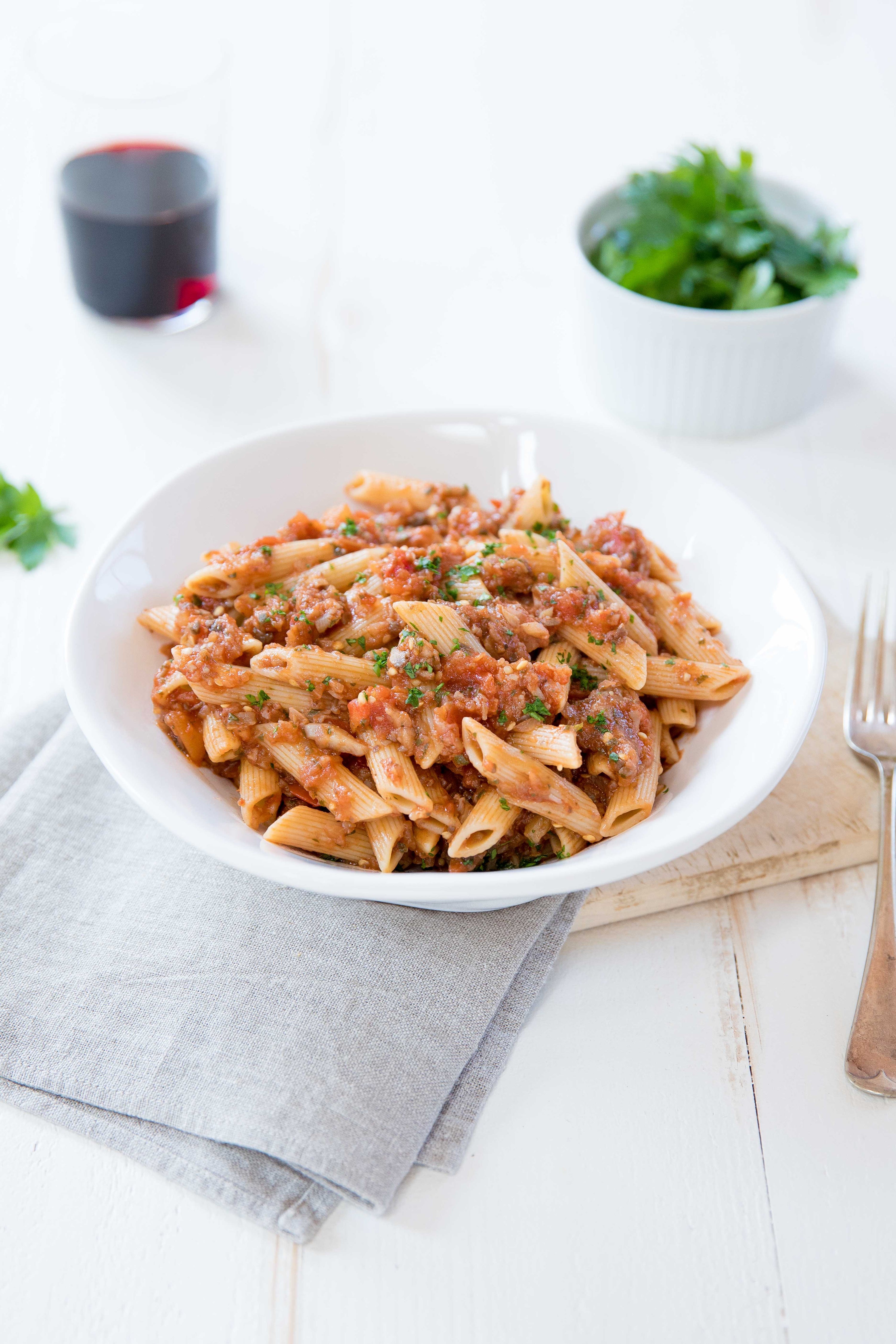 Penne pasta with eggplant and mushroom tomato sauce in a white bowl on a light wooden table with napkin and parsley