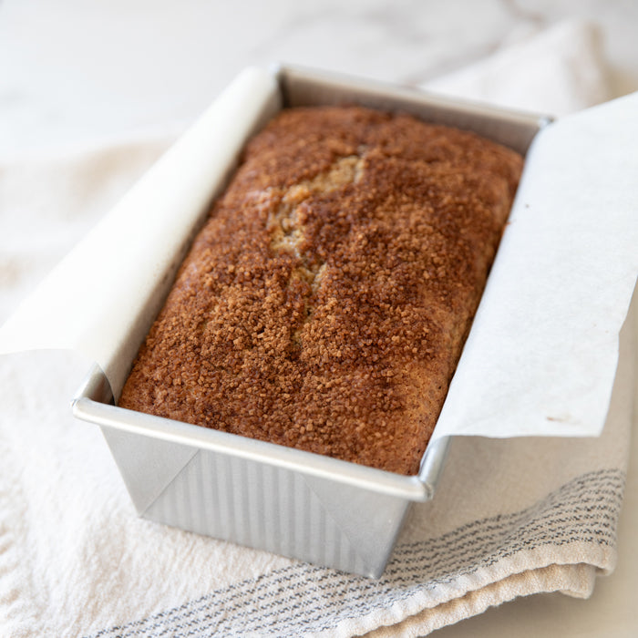 Angled top view of Einkorn apple cider loaf cake with coarse turbinado sugar crust in parchment-lined pan on striped towel
