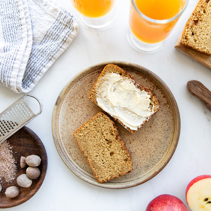Einkorn apple cider loaf cake slices on a plate, one slice spread with butter, served with apple halves, nutmeg, and glasses of apple cider