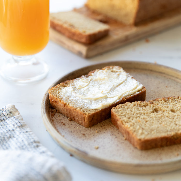 Sliced Einkorn apple cider loaf cake on a plate with one buttered slice and a glass of cider in the background