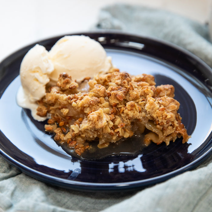 Close-up of a slice of Crunchy Einkorn apple crumble with oat and nut topping and vanilla ice cream on a dark blue plate