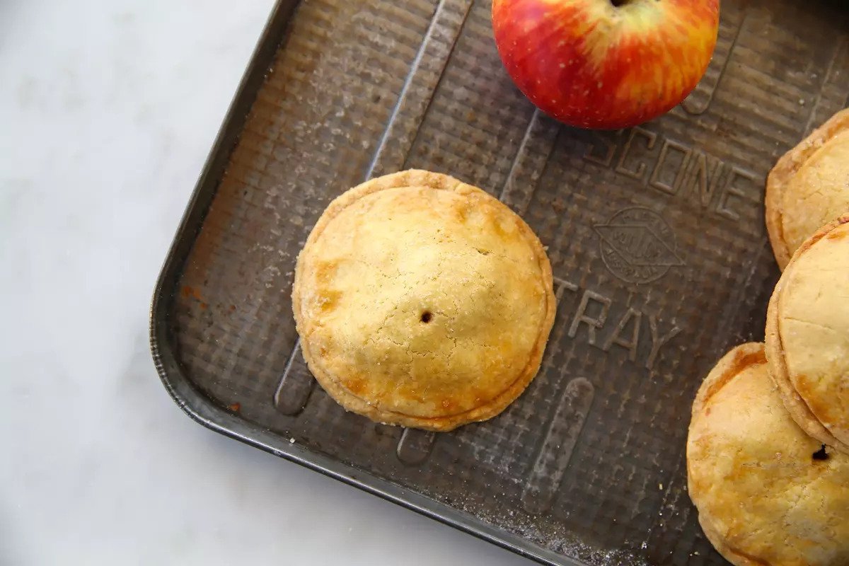 Top-down view of baked Einkorn apple hand pies with golden flaky crust on a dark baking tray and a whole apple