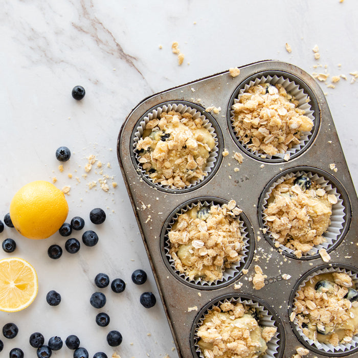 Einkorn blueberry streusel muffins in a muffin tin with scattered blueberries and a lemon on a marble surface