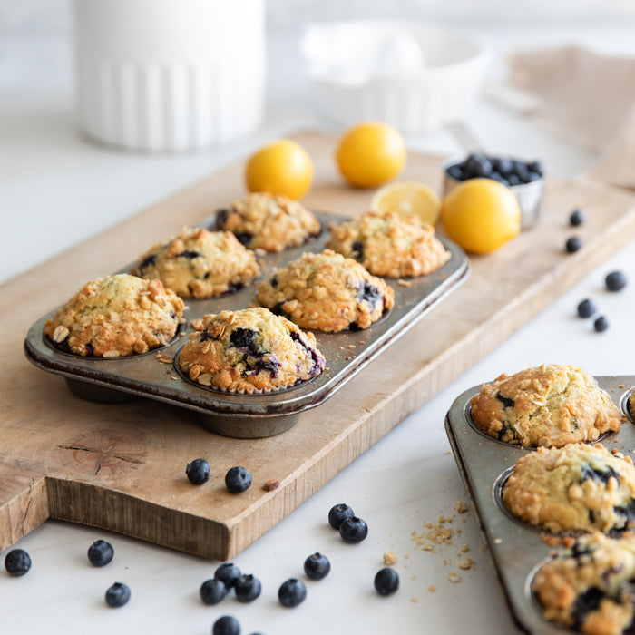 Blueberry streusel muffins made with einkorn cooling in muffin tins on a wooden board with scattered blueberries and lemons