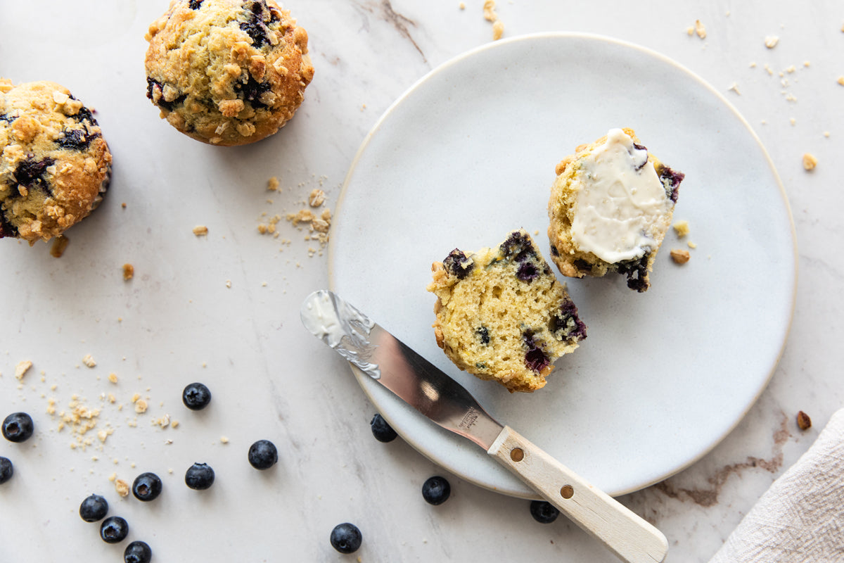 Einkorn blueberry streusel muffin halved and buttered on a plate with two whole muffins and scattered blueberries