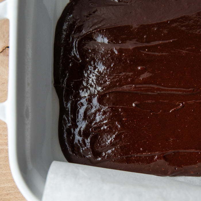 Close-up of Einkorn brookie batter spread in a parchment-lined baking dish showing a glossy chocolate surface