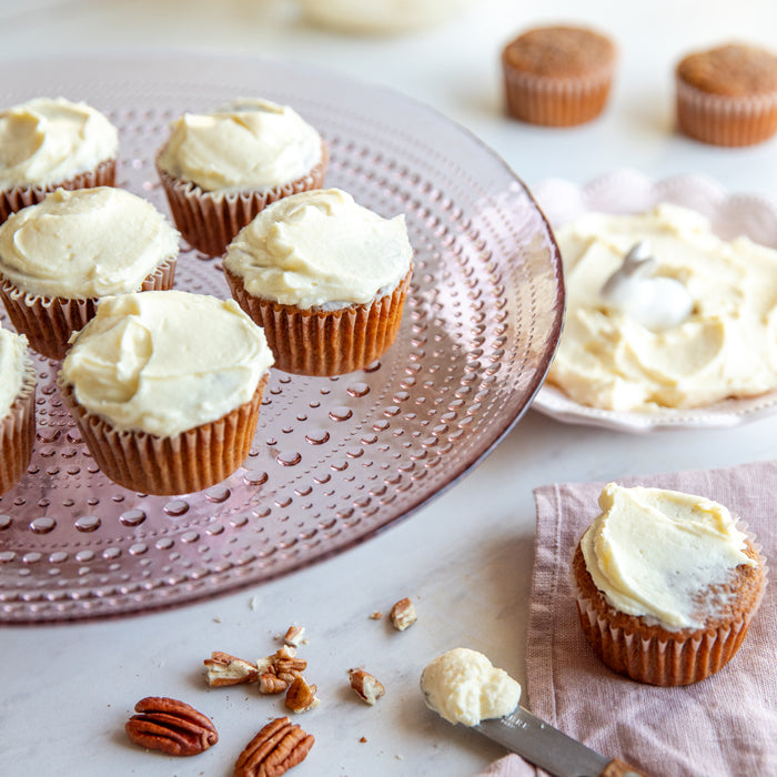 Einkorn carrot cupcakes topped with cream cheese frosting on a pink glass cake stand with extra frosting and pecans nearby