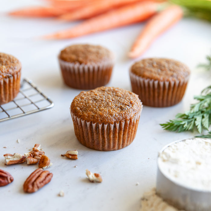 Three Einkorn carrot cupcakes arranged on a marble surface with pecans and a flour sifter, carrots blurred in the background