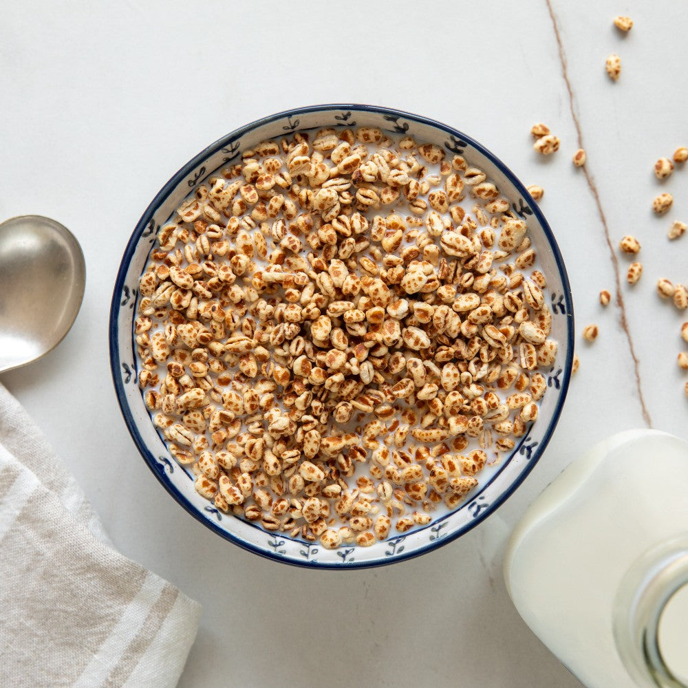 Bowl of Organic Einkorn cereal puffs with milk on a light tabletop beside a spoon and a glass milk pitcher