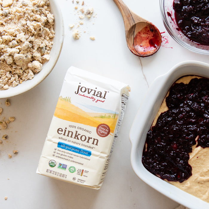 Overhead of Jovial Einkorn flour bag with pistachio streusel, wooden spoon, and cherry-filled baking dish