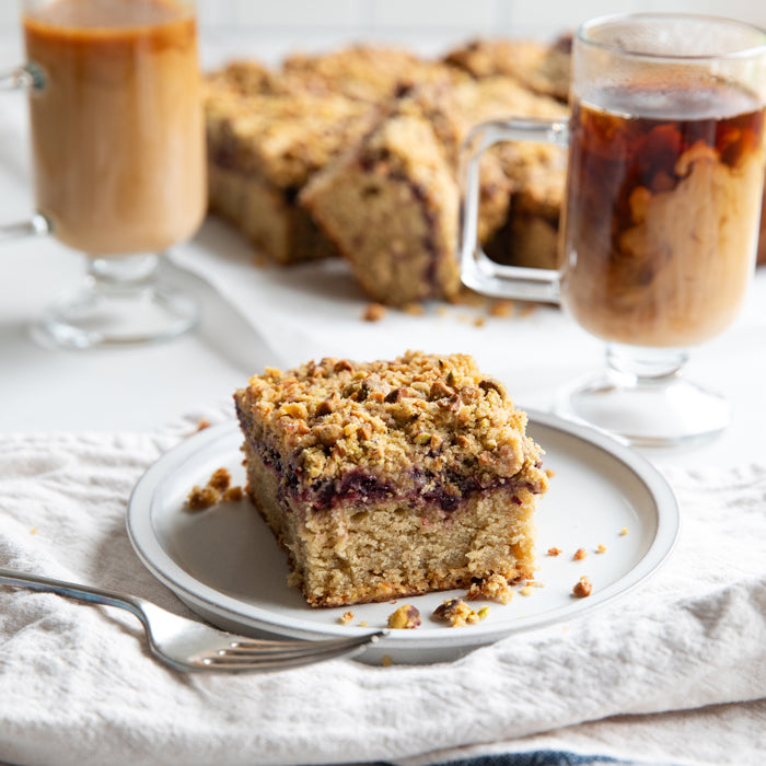 Close-up of a slice of einkorn cherry coffee cake with cherry jam and pistachio streusel on a plate, coffee in background