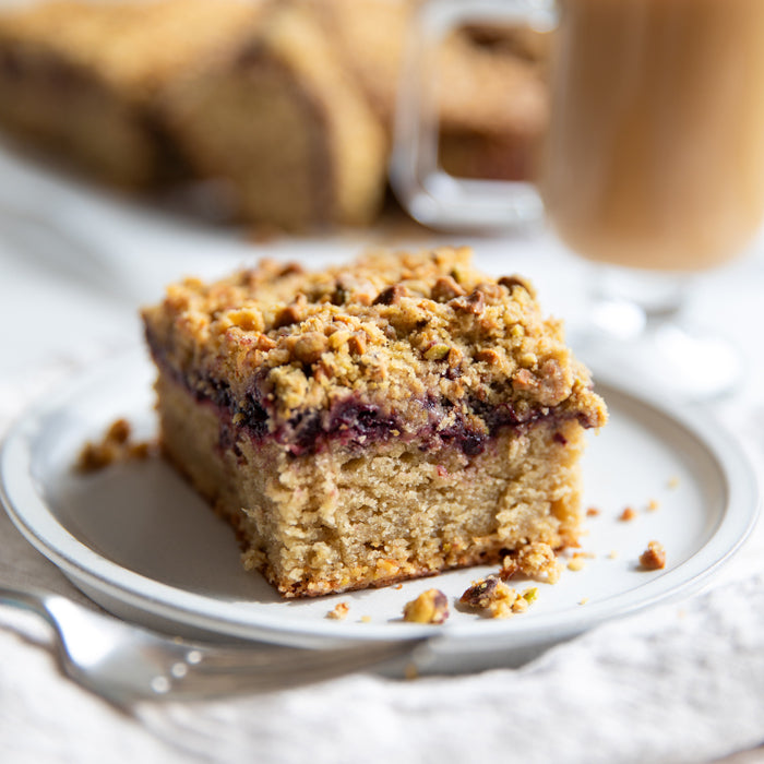 Close-up of a slice of Einkorn cherry coffee cake showing a cherry jam layer and pistachio streusel on a white plate